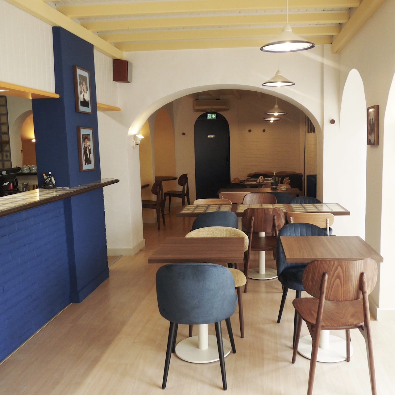 Vue profonde de la première salle de la crêperie avec poutres soulignées en jaune pâle et le bloc bleu du bar visible.